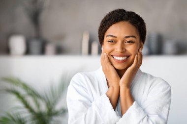 Beautiful Black Young Woman Wearing White Bathrobe Standing In Stylish Bathroom Interior, Happy African American Female Touching Face And Smiling, Enjoying Beauty Treatments And Skincare, Copy Space