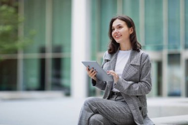 Cheerful young businesswoman working on tablet computer while sitting outdoors, websurfing near business center, looking aside with smile. Modern professional life, internet and gadgets