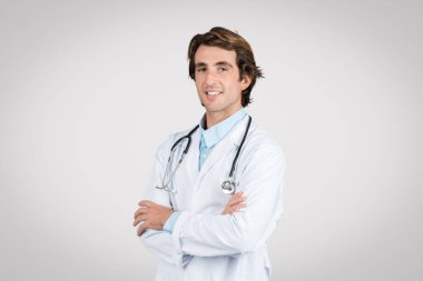 Smiling young european male doctor in white coat with stethoscope around his neck standing confidently with arms crossed against gray background