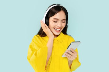 Happy young brunette lady watching video, listening to favorite music or podcast on cell phone, using wireless headphones, blue studio background. Modern gadgets and entertainment, spare time