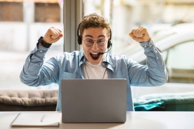 Glad young european guy in glasses, headphones using laptop, rises fists up, celebrate win, success at remote work or online communication in cafe. Winner gesture, game