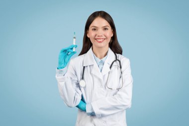 Young, professional woman doctor in white coat and gloves ready with syringe, standing against simple blue background for medical imagery.