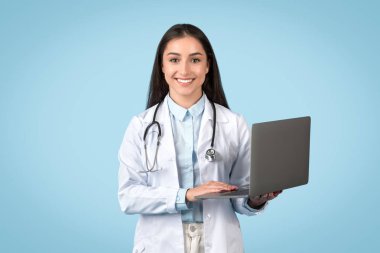 Professional female doctor holding laptop, representing telemedicine services, with friendly smile and wearing white lab coat, blue background