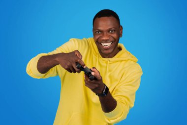 A joyful happy Black man gamer in a yellow hoodie wields a game controller with dynamic enthusiasm, showcasing the fun of gaming against a vivid blue studio background