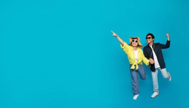 Ecstatic millennial Asian couple in summer attire are dancing and pointing joyfully, exuding happiness and carefree spirit on a bright turquoise background with ample copy space