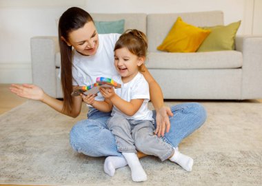 Mother and little child laughing while playing with toy at home, happy mom and her toddler daughter sharing joyful moment, having fun together on floor in cozy living room interior, free space