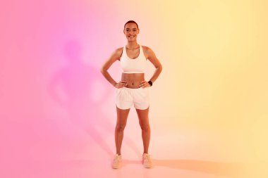 A confident female athlete stands with hands on hips in white sportswear, her shadow cast on a vibrant pink to yellow gradient background, exuding health and fitness. Sport, fitness and weight loss