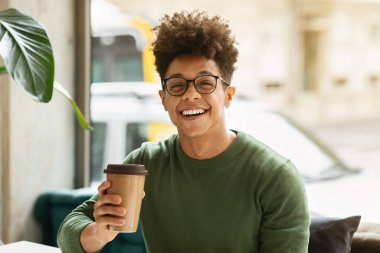 Portrait of happy stylish young black guy wearing eyeglasses enjoying coffee at cafe, holding paper cup and smiling at camera. Millennials lifestyle, leisure concept