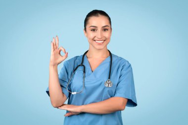 Female nurse in cheerful mood, smiles at the camera, and makes an OK gesture with her hand against blue background, conveying positivity in healthcare