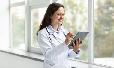 Modern healthcare. Cheerful beautiful long-haired young woman wearing medical white coat uniform doctor standing by window at clinic, using digital tablet, chatting with client, copy space
