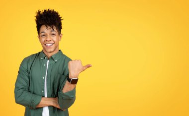 Cool handsome stylish millennial african american guy pointing at copy space with thumb, showing mockup for advertisement, isolated on yellow studio background