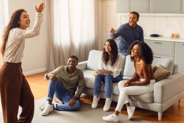 Young international students playing charades in the living room, laughing enjoying home party together. Group of multiethnic friends engaged in game of pantomime. Friendship and Entertainment