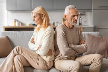 Upset senior married couple grey-haired man and blonde woman sit back to back on couch at home, not talk to each other, feeling down after fight, have difficulties in marriage, thinking about divorce