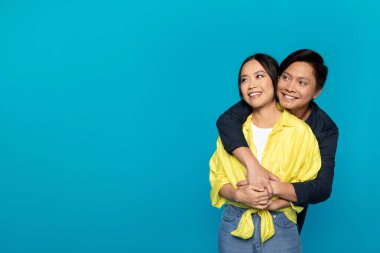 Smiling Asian couple in casual clothing embracing fondly, man hugging woman from behind, both looking hopeful and happy against a bright turquoise background with ample copy space