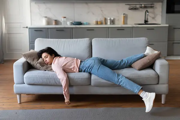 Woman in pink blouse and jeans lying exhausted on couch, taking well-deserved rest after completing her household cleaning tasks