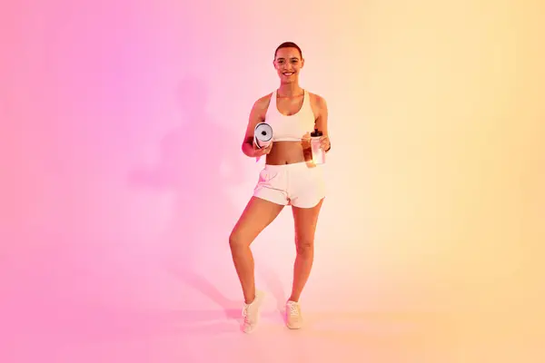 A smiling latin woman with a shaved head holds a yoga mat and a water bottle, ready for her fitness routine, in white sportswear against a pink and yellow gradient backdrop