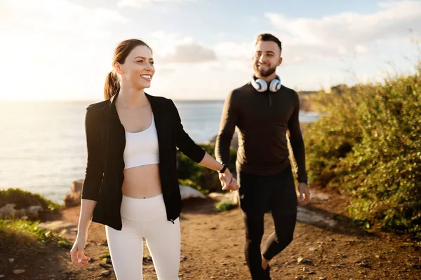 Cheerful man and woman in stylish fitness attire holding hands and walking together along a scenic coastal path, exuding health and happiness