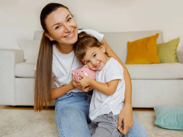 Wise Economy. Happy Mother And Little Daughter Holding Pink Piggy Bank, Smiling Young Mom And Cute Toddler Female Child Saving Money For Future, Sitting Together On Floor In Living Room At Home