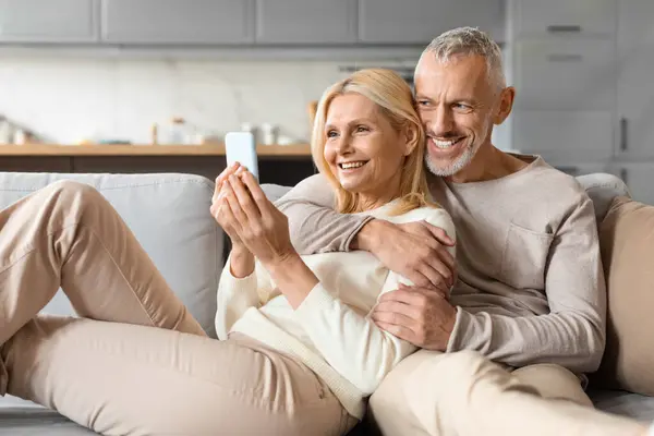 Happily married senior couple sitting together on sofa at home, using smartphone, websurfing, scrolling. Cheerful loving man and woman spouses shopping online, taking selfie, copy space