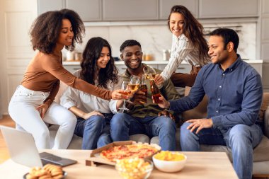Diverse group of students clinking with bottles and glasses of beer, sharing stories and laughs in a modern living room setting, enjoying party gathering with pizza, sitting on sofa indoors