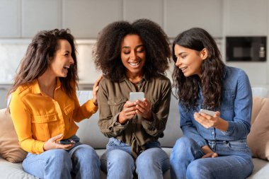 Three multiethnic women friends websurfing on cell phones while sitting on couch and laughing, looking at smartphone watching funny videos together, scrolling social media app in living room
