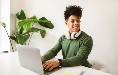 Happy stylish millennial black man with wireless headphones using laptop, sitting at cozy cafe, looking aside at copy space and smiling. Freelancer working on computer, websurfing