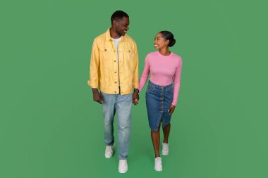 Happy millennial black man and woman wearing casual outfit have date, holding hands and walking, enjoying conversation, time together, isolated on green studio background, full length