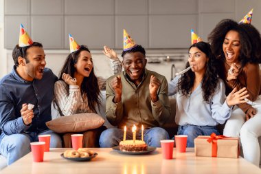 Happy multiracial students celebrating birthday of black guy, cheering while he makes wish and blowing candles on cake in modern kitchen interior. Bday party with true friends