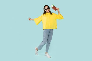 Vacation, traveling, hot offer. Positive stylish young woman wearing sunglasses with passport and flight tickets in her hand dancing over blue studio background, copy space, full length