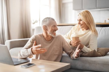 Cheerful senior couple using computer laptop at home, sit on couch in cozy interior, drinking tea, planning weekend or holiday, booking tickets and hotel on Internet, have conversation, copy space