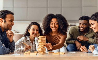 Young diverse friends gathered around living room table indoor, engaged in board game building wooden tower together, showcasing the fun of their multicultural friendship