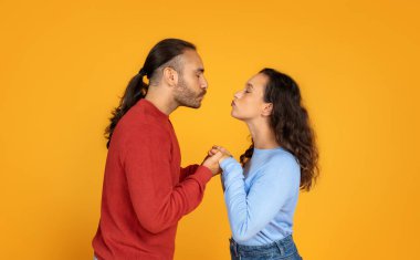 Romantic millennial man and woman holding hands and kissing on orange studio background. Young european couple have date, cuddling, celebrating anniversary or St. Valentines Day