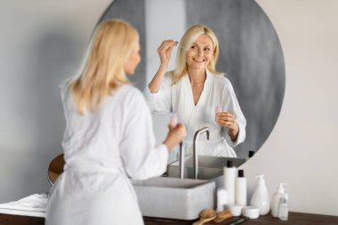 Elegant senior woman in white bathrobe applying facial serum, looking at her reflection in bathroom mirror, happy smiling elderly female enjoying her beauty routine at home, copy space