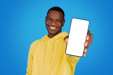 Cheerful african american man student blogger in a bright yellow hoodie holding a smartphone with a blank screen towards the camera on a blue studio background, recommendation app
