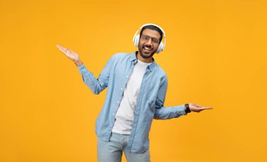 An exuberant man in a denim shirt wears white headphones and gestures with his hands, showing excitement or dancing to music, against a vibrant orange background