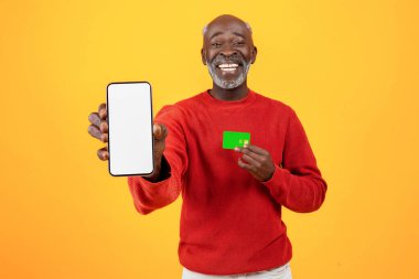 Smiling senior african american man show credit card and smartphone with empty screen, recommend banking app on device, isolated on orange studio background. Shopping, sale and money