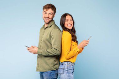 Happy young couple standing back-to-back, each using their own smartphone with joyful expressions, symbolizing individual digital engagement in social setting, against blue background