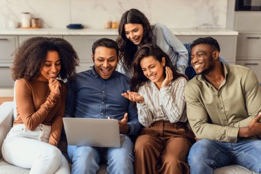 Group of young multiethnic friends watching funny comedy movie on laptop gathered in modern living room indoor. Fellow students enjoying weekend browsing and watch videos online