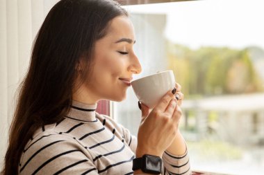 Glad young caucasian lady with smartwatch enjoy coffee cup, sunlit window, embodying relaxed lifestyle at home, close up. Tasty fresh drink at spare time at morning, break