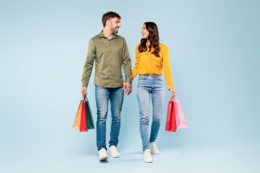 Relaxed and happy couple enjoying shopping trip, holding hands and carrying colorful bags, embodying carefree and contented retail experience against soothing blue background