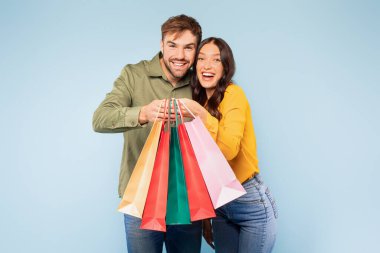 Radiant young couple holding collection of colorful shopping bags, their faces beaming with the thrill of successful shopping day, set against bright blue background