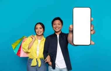 Happy Asian couple with colorful shopping bags and a modern smartphone, presenting a blank phone screen with excitement against a vibrant blue backdrop, ideal for advertising