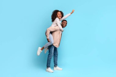 Ecstatic black woman riding piggyback on cheerful man, both with joyous expressions and woman raising her arm in victory, against light blue background, full length