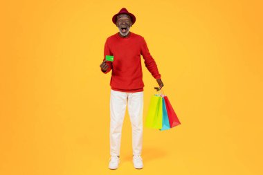 Glad surprised senior african american man shopaholic in hat with open mouth and many bags with purchases, show credit card, isolated on orange studio background. Shopping, sale and money