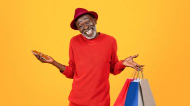 Smiling confused old black man shopaholic in hat, shrugs shoulders with many bags and phone, isolated on orange studio background, panorama. Shopping online app, huge sale and choose