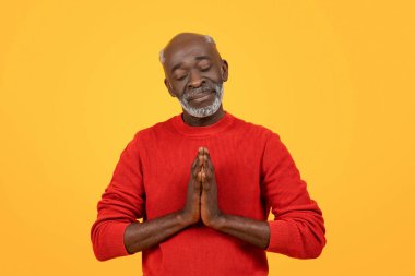 Peaceful senior african american man with a white beard in a meditative pose with hands together, eyes closed, wearing a red sweater against a calming yellow studio background