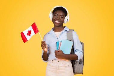 Cheerful millennial black lady student in wireless headphones, with books, flag of Canada, listen lesson, isolated on yellow studio background. Study and education exchange, learn French, English