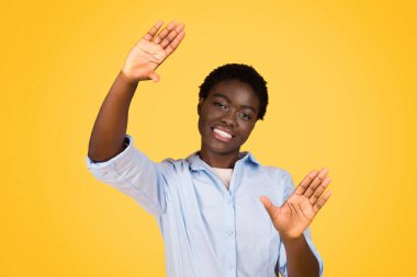 A black woman student playfully making a frame sign with her hands, suggesting creativity and vision, her joyful demeanor set against yellow background, capturing a moment of imaginative expression