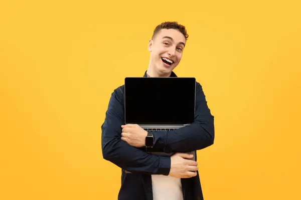 Modern gadgets and technologies. Happy young guy hugging his brand new laptop with blank black screen and smiling at camera, posing isolated on yellow studio background