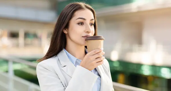 Focused caucasian businesswoman sipping coffee from a takeaway cup, dressed in a professional light beige blazer over a blue shirt, contemplating her next meeting, panorama